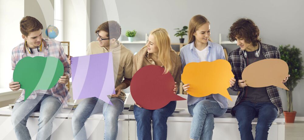 Jugendbeirat Partnerschaft für Demokratie Landkreis Görlitz Fünf Jugendliche sitzen in einer Reihe auf zwei aneinandergereihten Schreibtisches und halten unbeschriftete Buntpapier in verschiedenen Formen als eine Sprechblase in den Händen u. blicken mit einem lachendem Gesicht auf die Sprechblasen des Sitznachbarn