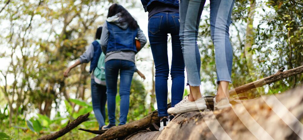 Jugendliche balancieren auf einen am Boden liegenden Baumstamm einen Waldpfad entlang.